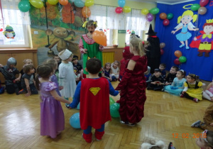 Nauczycielka w przebraniu z balonem w ręku, przy niej pięcioro dzieci tańczy w kole z balonami. Pozostali kibicują.